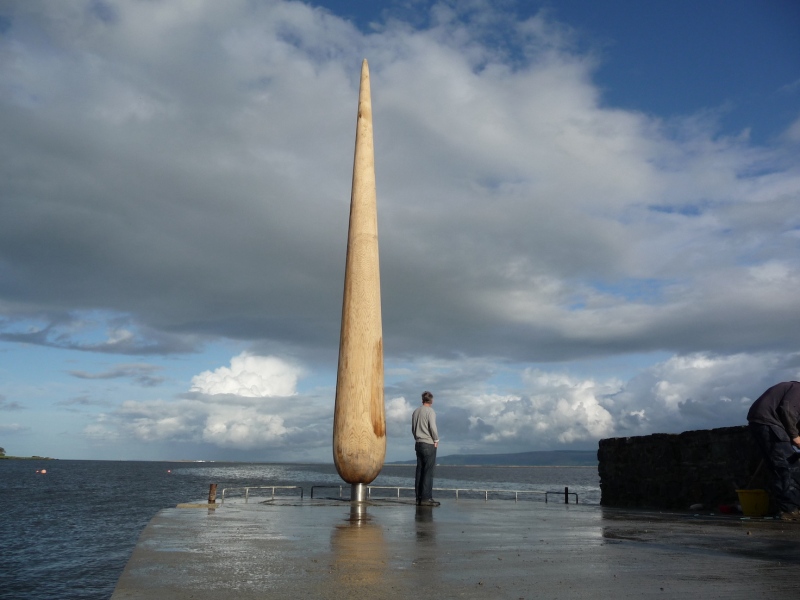 The Fid (2007) Oak, stainless steel. Old Stone Pier, Moville, Co. Donegal. 'This scene of Moville&rsquo;s historic Old Stone Pier, linking the River Foyle with the land of Inishowen, now features the striking addition of a new sculpture by artist Locky Morris (carved from a single trunk of oak) This commission from Donegal County Council Public Art Office follows on from the artist&rsquo;s commission for Pole Star, his large sculpture which has become an iconic feature for Letterkenny. The Fid at Moville Old Stone Pier has its inspirations within the village&rsquo;s powerful connections to the sea and seafaring. The artist&rsquo;s conversations with local people uncovered tales of the beacon fires, which were once lit on the hills of Inishowen to mark the passage down the River Foyle of emigration ships. And in Greencastle Maritime Museum the artist encountered an example of a &lsquo;fid&rsquo;, the small handheld tool turned from a piece of wood, used by fishermen for splicing ropes and nets and by sailmakers for opening holes in the sails without tearing the material. The final sculpture both recollects those beacon flames and that form of a &lsquo;fid&rsquo;. It unites this elegant and flowing form with an assured sense of scale and location &ndash; standing over 25 feet high and over three feet wide at base, sited at the end of the pier, facing directly onto a beautiful and dramatic seascape. The Old Pier then, marks the village&rsquo;s maritime heritage, for individual seafarers, emigrants and immigrants, their families, and the wider communities. In many ways, The Fid itself will act as a beacon towards this location: concentrating our vision and our thoughts upon those links between sea and land, which have made Moville'