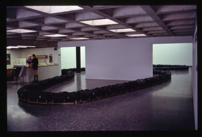 Aristocrat (1991) gallery installation. Large holdall bags (polyester/nylon), teak veneered chipboard, furniture latches, furniture castors &nbsp;(installation view, Mannheimer Kunstverein, Germany) '...In 1991 Morris was invited to make an artwork at the Mannheim Kunstverein in Germany. The space was large reminding him of an airport lounge. It was also not long after the Berlin wall had come down and there was free movement of people from the former east Germany section. Indeed in the new Europe, as an economic block, cheap labour would be on the move with more urgency. And Mannheim itself was the centre of strategic military planning during the Gulf War. Even the scale and quantity of heavy articulated lorries on the German Autobahns seem to the artist to reinforce the tension in the air.&nbsp; In response to the above factors and the scale and ambience of the gallery space Morris made a floor piece called&nbsp;Aristocrat.&nbsp;Like&nbsp;Cortege&nbsp;this installation snaked its way around two rooms. The work consisted of a series of cheap travelling bags (Aristocrat being their trade name) placed on wooden bases with wheels. It marks an important transition for the artist: increased scale; more minimalist in expression but not in concept and less representational in appearance. It was an experimental extension of means...'&nbsp;&nbsp; (text extract, The City as Art, Liam Kelly, A.I.C.A , Irish section, 1994)